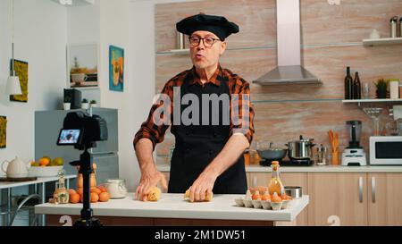 Nonno che registra video di cibo in cucina, preparando pizza fatta in casa. Pensionato blogger chef influencer utilizzando la tecnologia di comunicazione Internet, tiro blogging sui social media con le apparecchiature digitali Foto Stock