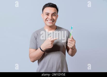 Bel ragazzo che tiene e indica lo spazzolino isolato. Persona che tiene e punta uno spazzolino da denti su sfondo isolato, sorridente uomo che tiene e poi Foto Stock