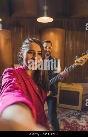 Happy Brown-haired woman vocalist prende un selfie con un chitarrista maschile. Pratica rock band prima del concerto. Ripresa verticale per interni. Foto di alta qualità Foto Stock