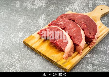 Una vista dall'alto di bistecche di picanha crude su un tagliere di legno e un tavolo grigio Foto Stock