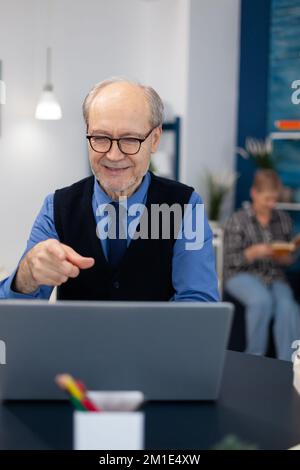 Un uomo anziano felice che punta al laptop durante la videoconferenza. Pensionato allegro con capelli grigi che sorridono alla webcam del computer portatile mentre chiacchiera seduto alla scrivania. Foto Stock