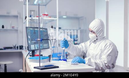 Medic indossa tuta dpi in caso di epidemia globale lavorando con micropipetta in fiasche di vetro. Chimico in laboratorio moderno che fa ricerca utilizzando dispenser durante l'epidemia globale con COVID-19. Foto Stock