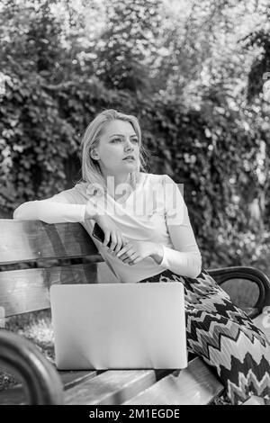 Creazione di nuovi contenuti per il suo blog. Ragazza blogger funziona all'aperto con computer portatile e smartphone. Donna lavora su blog web nel parco. Ragazza freelancer riuscito Foto Stock