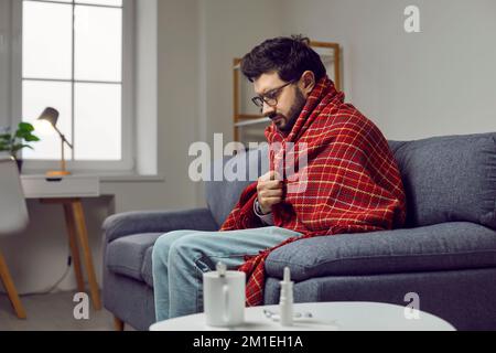 Giovane ammalato in una coperta seduta sul divano a casa, sentendosi freddo e brivido Foto Stock