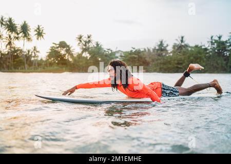 Uomo nero dai capelli lunghi che si paddling su una lunga tavola da surf al punto di surf nell'oceano Indiano. Palmeto ha littato raggi del tramonto sullo sfondo. Acqua estrema s Foto Stock