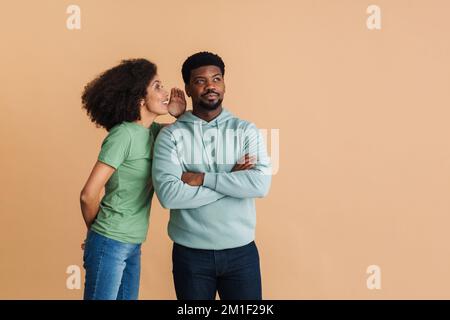 Donna felice nera sorridente e sussurrante segreto all'orecchio dell'uomo isolato su sfondo beige Foto Stock