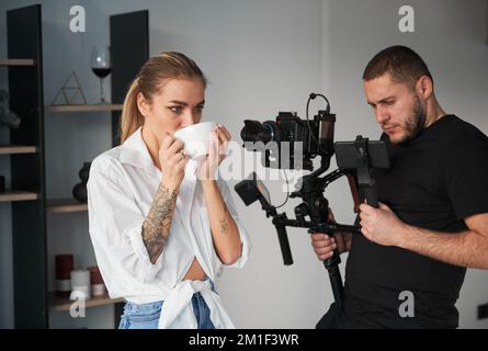 Uomo videografo che scatta riprese con la modella femminile, utilizzando una fotocamera montata su un'apparecchiatura di stabilizzazione del gimbal all'interno. Giovane donna con tatuaggi sulle mani che posano e bevono tè caldo. Foto Stock