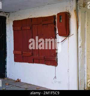 Una finestra rossa in legno e una cassetta del contatore elettrico su una vecchia parete bianca. Foto Stock