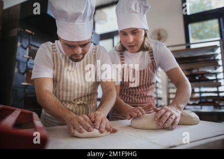 Uomo con giù sindromi aiutare a preabbinare il pane in panetteria con il suo collega. Concetto di integrazione delle persone con disabilità nella società. Foto Stock