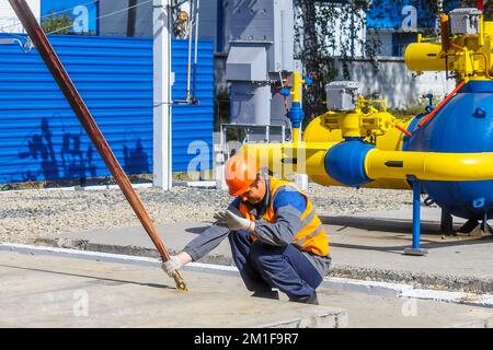 Slinger posa la lastra di cemento in cantiere il giorno estivo. Il lavoratore in giubbotto di protezione e casco da costruzione supervisiona la posa della base sul cantiere. Foto Stock