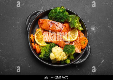 Pesce sano al forno bistecche di salmone, broccoli, cavolfiore, carota in casseruola in ghisa su fondo di pietra nera scura. Cucinare un delizioso basso Foto Stock