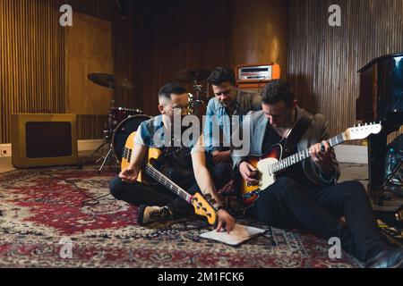 due giovani chitarristi e un batterista seduti sul pavimento e guardando il foglio di note sulla ripetizione, registrando il concetto di musica da studio. Foto di alta qualità Foto Stock