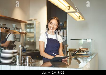 Ritratto di bella ragazza asiatica sorridente, barista in caffetteria che lavora dietro il bancone, utilizzando il tablet come terminale POS, l'elaborazione dell'ordine Foto Stock