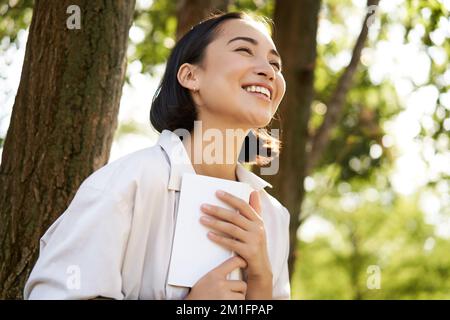 Romantico sorridente ragazza lettura libro in parco o foret, seduta sotto l'ombra dell'albero nella giornata di sole, relax all'aria fresca circondata dalla natura Foto Stock