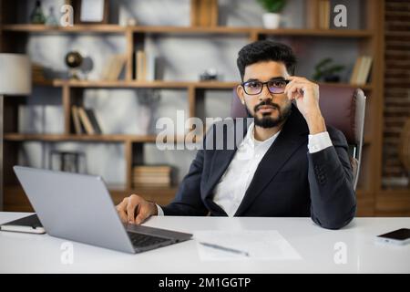 Giovane uomo d'affari bearded che usa il computer portatile durante la videochiamata che lavora in ufficio. Adulto concentrato uomo di successo con vestito ufficiale seduto al tavolo di legno al coperto. Foto Stock