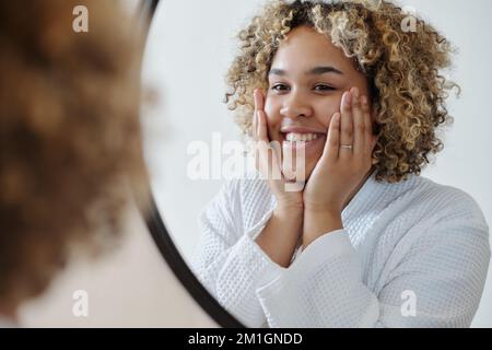 Giovane donna felice e sana in accappatoio bianco in piedi di fronte allo specchio e prendersi cura del suo viso mentre godendo l'igiene del mattino Foto Stock