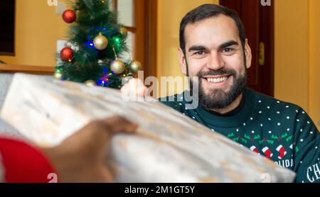 Felice uomo bearded sorprendente con un regalo avvolto di fronte a un albero di natale che indossa un maglione di natale Foto Stock