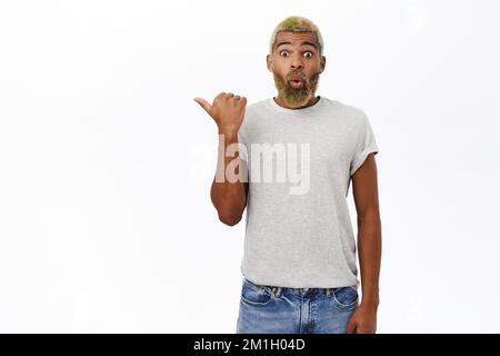 Ragazzo impressionato con la barba gialla, l'uomo afroamericano che punta a sinistra, dicendo WOW, mostrando la pubblicità, quel gesto di senso, sfondo bianco Foto Stock