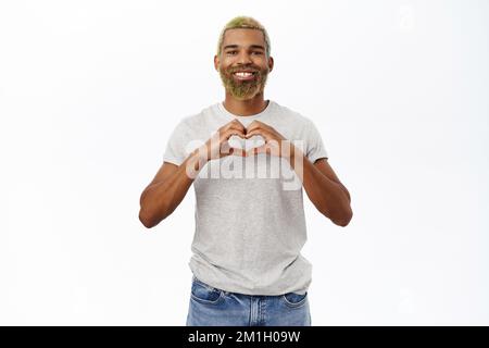 Sorridente bel ragazzo mostra amore e cura gesto, rende mano cuore e guarda come la macchina fotografica, si alza su sfondo bianco Foto Stock