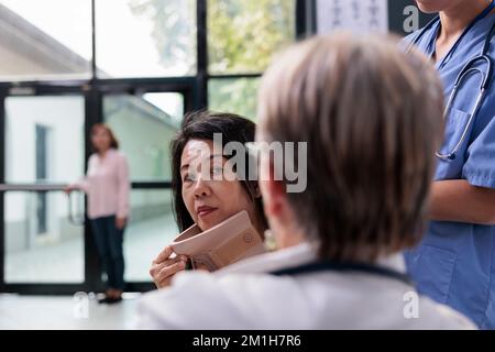 Specialista di salute aiutando la donna anziana a togliere il colletto, cercando di recuperare dopo l'incidente. Paziente ferito con supporto cervicale in schiuma che riceve aiuto da medico e assistente nella lobby dell'ospedale Foto Stock