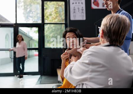 Medico traumatologo e infermiere che aiutano il paziente anziano a togliere il colletto cervicale durante l'appuntamento per l'esame nell'area di attesa dell'ospedale. Donna asiatica che riceve assistenza medica Foto Stock