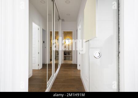 Un lungo corridoio in una casa con materiali bianchi alle pareti, porte in legno e un armadio con porte a specchio scorrevoli Foto Stock