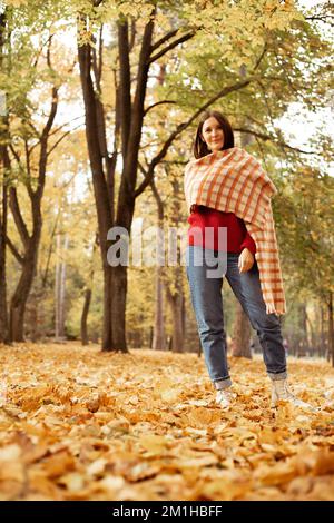 Ritratto di giovane donna attraente in piedi vicino agli alberi tra foglie gialle cadute nel parco forestale. Verticale. Autunno. Foto Stock