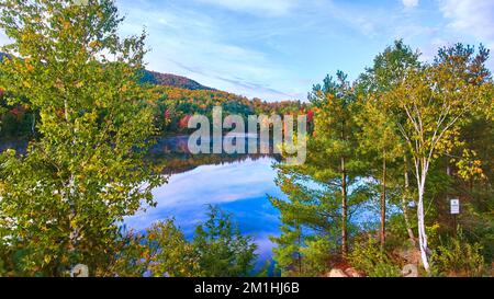 Guardando attraverso gli alberi vista elevata del lago mattina nebbia blu circondato da montagne di caduta Foto Stock
