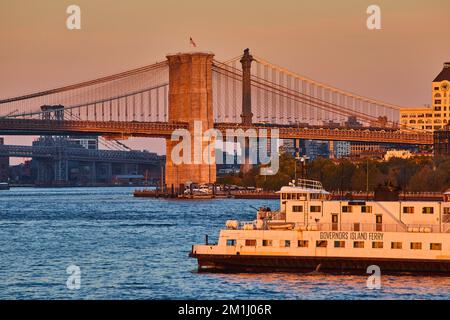 Traghetto per Governors Island in un'ora d'oro al tramonto, luce dalle acque che passano dal Ponte di Brooklyn con bandiera americana Foto Stock