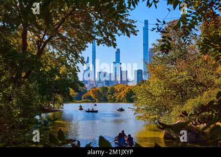 Vista attraverso gli alberi di stagno con le barche a Central Park New York City con grattacieli sullo sfondo Foto Stock
