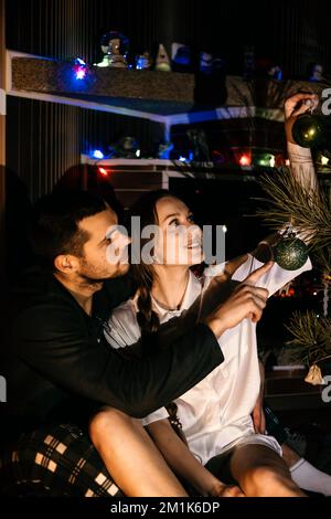 Buon Natale. Giovane coppia innamorata che festeggia il Natale o la vigilia di Capodanno a casa. Coppia romantica in pigiama seduta e abbracciata vicino a natale Foto Stock