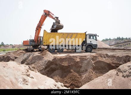 l'escavatore cingolato arancione versa la terra nel cassone del dumper giallo. Momento di lavoro in cantiere in lavorazione di terracotta. Caricamento e trasporto Foto Stock