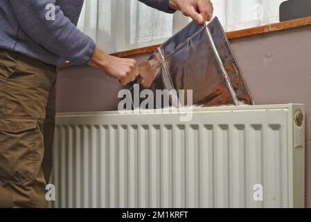 Uomo che installa pannelli in lamina isolante riflettente dietro un radiatore per migliorare l'efficienza del riscaldamento e cercare di risparmiare denaro sulle bollette utilizzando meno energia. Foto Stock