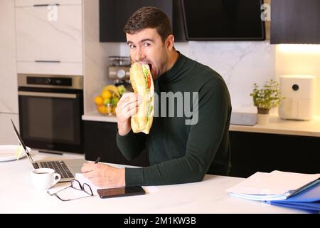 Lavoratore che mangia un panino molto grande in ufficio domestico Foto Stock