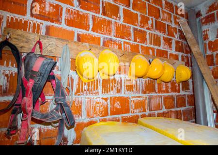 Le attrezzature di lavoro industriali dei lavoratori, i caschi gialli e le cinture di sicurezza sono appesi su un appendiabiti realizzato localmente in legno e chiodi, fissato su mattoni rossi W. Foto Stock