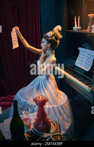 Giovane tenera ragazza a immagine di una persona medievale in elegante vestito bianco seduto al pianoforte e imparando una nuova composizione musicale. Confronto delle ere Foto Stock