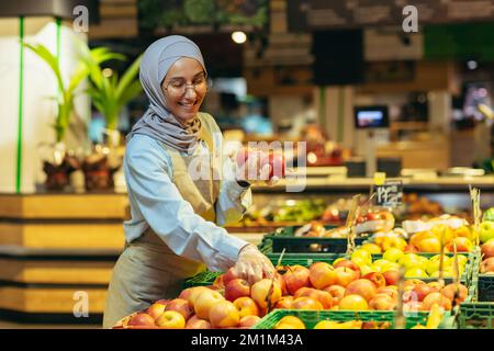Venditore femminile in hijab navigazione e controllare le mele in supermercato, donna in grembiule sorridente al lavoro in negozio nel reparto frutta e verdura. Foto Stock