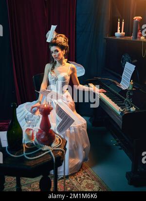 Caos creativo. Ritratto di bella giovane donna a immagine di medievale in tenero vestito bianco seduto al pianoforte e suonando. Confronto delle ere Foto Stock