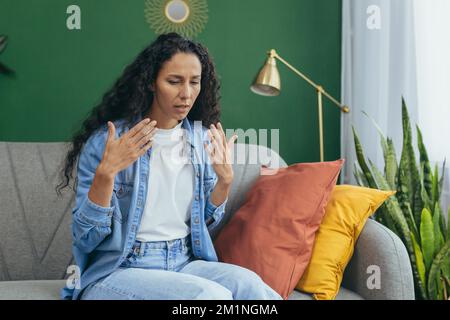 La donna è calda nella stanza, la donna ispanica seduta sul divano e che agita le mani cercando di rinfrescarsi e raffreddare l'aria a casa nel soggiorno. Foto Stock