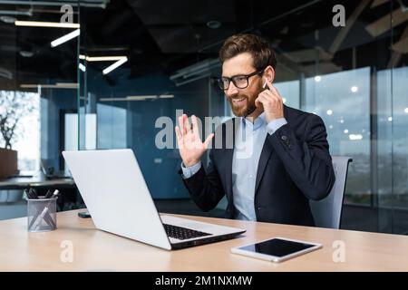 Incontro online con i clienti, conferenza. Un giovane uomo d'affari in un vestito siede in ufficio al tavolo. Parla durante una videochiamata da un computer portatile. Tiene l'auricolare in mano, dice ciao, sorride. Foto Stock