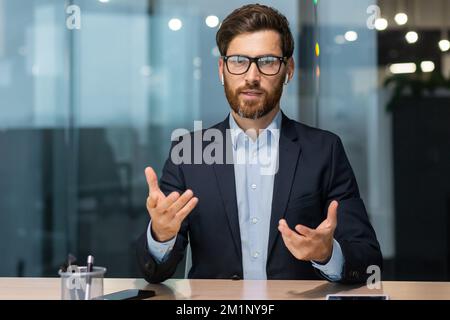 Un giovane uomo d'affari bello conduce la formazione di affari in linea. Si siede in ufficio di fronte alla telecamera, insegna, consulta. Foto Stock