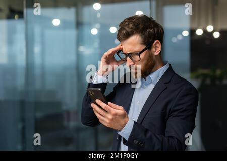 Un giovane uomo d'affari preoccupato tiene il telefono nelle sue mani, guarda. Lettura di cattive notizie, ricevuto un messaggio. Scioccato, tiene la testa. Foto Stock