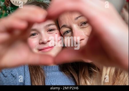 La famiglia amorevole trascorre le vacanze invernali a casa. Ritratto di felice sorridente bella madre e bambino che si uniscono le mani facendo forma di cuore seduto in una stanza festosa e accogliente con albero di Natale . Foto Stock