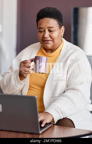 Ritratto verticale di donna nera anziana che usa un computer portatile a casa e che si diverte con una tazza di caffè Foto Stock