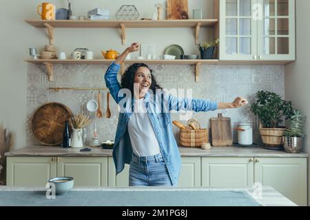 Giovane bella donna a casa cantando e ballando in cucina, allegra donna latino-americana che si diverte da sola con i capelli ricci. Foto Stock