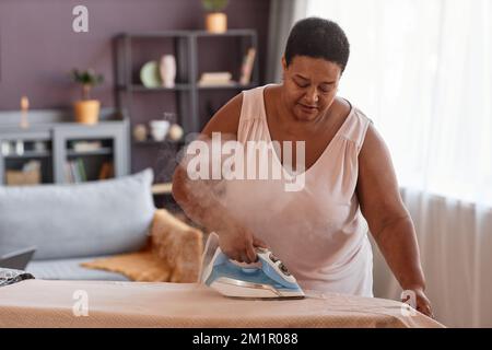 Vita in su ritratto di donna anziana nero stirare vestiti in un ambiente accogliente casa, spazio copia Foto Stock