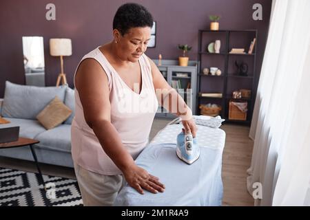 Vista ad alto angolo di una donna matura nera che stira i vestiti a casa mentre si prende cura della casa Foto Stock