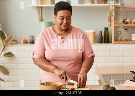 Vita in su ritratto di donna anziana nera che taglia le verdure mentre cucinando in cucina domestica accogliente, spazio della copia Foto Stock