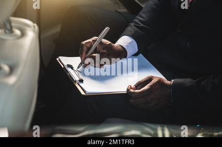 Concetto aziendale di un bell'uomo dalla pelle scura in tuta e occhiali nella seduta posteriore di un'auto di lusso che lavora e prende appunti. Firmare i documenti. chiudi Foto Stock