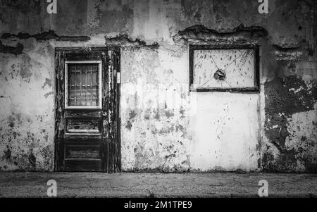 Una scala di grigi di una porta di legno intemperiata di un muro incrinato in un'area urbana - il concetto di povertà Foto Stock